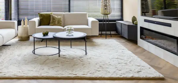 Ein heller, flauschiger Teppich liegt vor einem Sofa und einem Kamin. So wird der Teppich wieder flauschig gemacht.