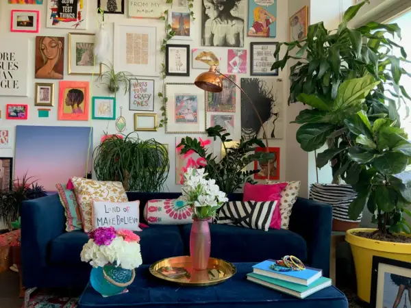 Ein Wohnzimmer im maximalistischen Stil mit einer blauen Couch, vielen Kissen, Pflanzen und einer Wand voller Kunstwerke und Drucke.