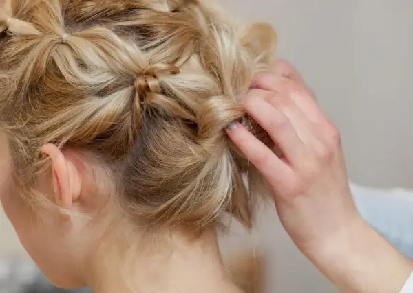 Eine Hand stylt blonde Haare zu verspielten hochsteckfrisuren mittellang. Kleine Zöpfe und gedrehte Strähnen ergeben einen lässigen Look.